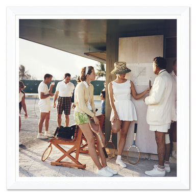 Vintage-style photo of people at a tennis court with a focus on a woman in a white dress and hat.
