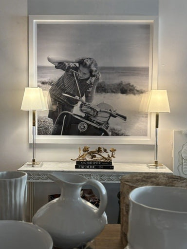 A large black and white framed art print depicting a woman on a vintage motorcycle by the beach, displayed on a console table in a styled room setting with two table lamps and ceramic vases.