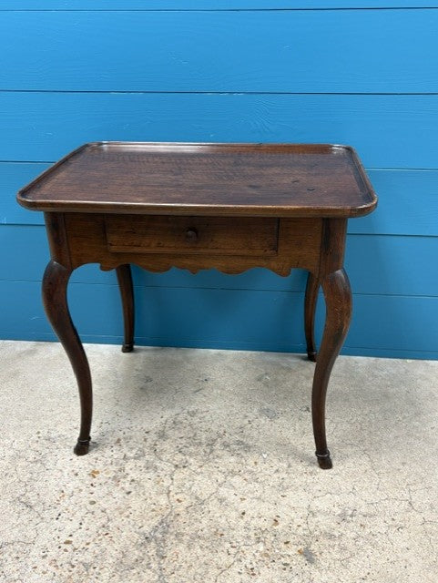 A dark brown wooden table with a single drawer and curved cabriole-style legs, positioned against a bright blue wooden wall on a concrete floor.