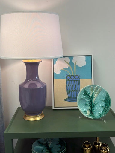 A purple ceramic table lamp with a white drum shade and gold base sits on a green side table next to a framed art print and a decorative teal plate.