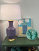 A purple ceramic table lamp with a white drum shade and gold base sits on a green side table next to a framed art print and a decorative teal plate.