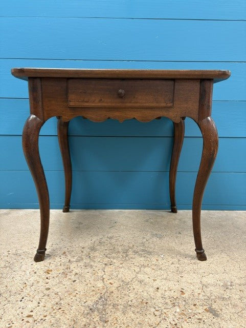 A vintage wooden table with a single drawer and curved cabriole-style legs, shown against a bright blue paneled wall and concrete floor.
