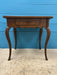 A vintage wooden table with a single drawer and curved cabriole-style legs, shown against a bright blue paneled wall and concrete floor.