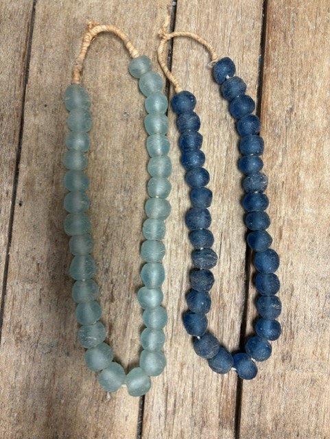 Two beaded necklaces made of round, matte beads, one in light blue-green and one in dark blue, hanging on a rustic wooden surface.