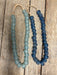 Two beaded necklaces made of round, matte beads, one in light blue-green and one in dark blue, hanging on a rustic wooden surface.