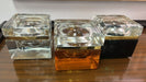 Three modern square glass lidded boxes, arranged side-by-side on a dark wooden table. features a gold metallic band and a black base.