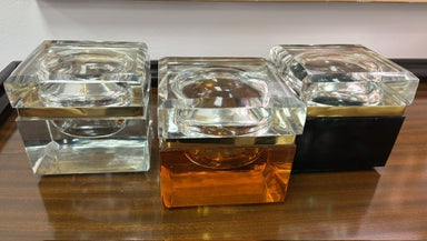 Three modern square glass lidded boxes, arranged side-by-side on a dark wooden table. features a gold metallic band and a black base.