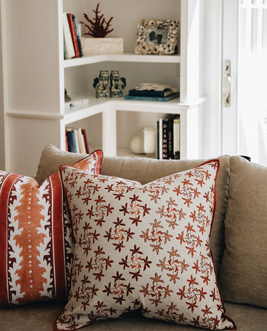 A decorative pillow with a floral pattern in brown and white colors, placed on a sofa with other pillows and a bookshelf in the background.