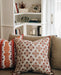 A decorative pillow with a floral pattern in brown and white colors, placed on a sofa with other pillows and a bookshelf in the background.