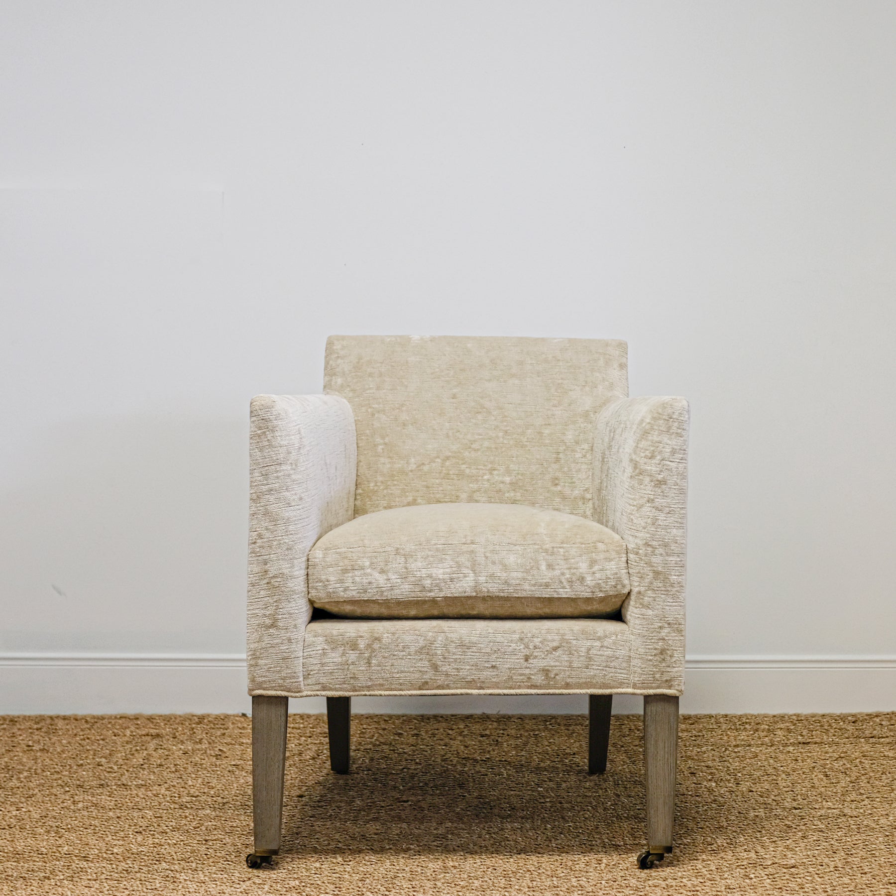 straight-on view of upholstered chair in mottled beige velvet with exposed wood leg, casters on front legs