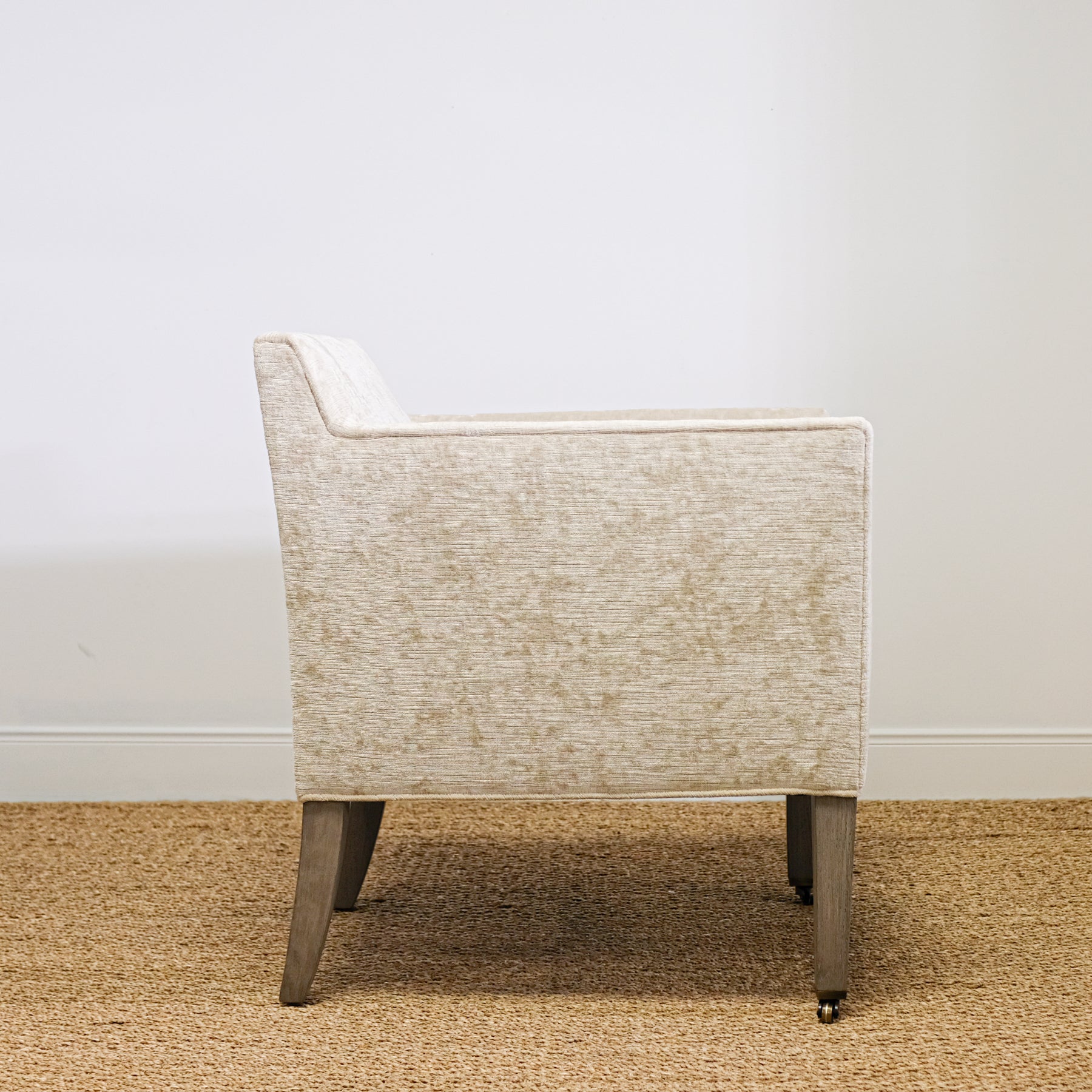 side view of upholstered chair in mottled beige velvet with exposed wood leg, casters on front legs