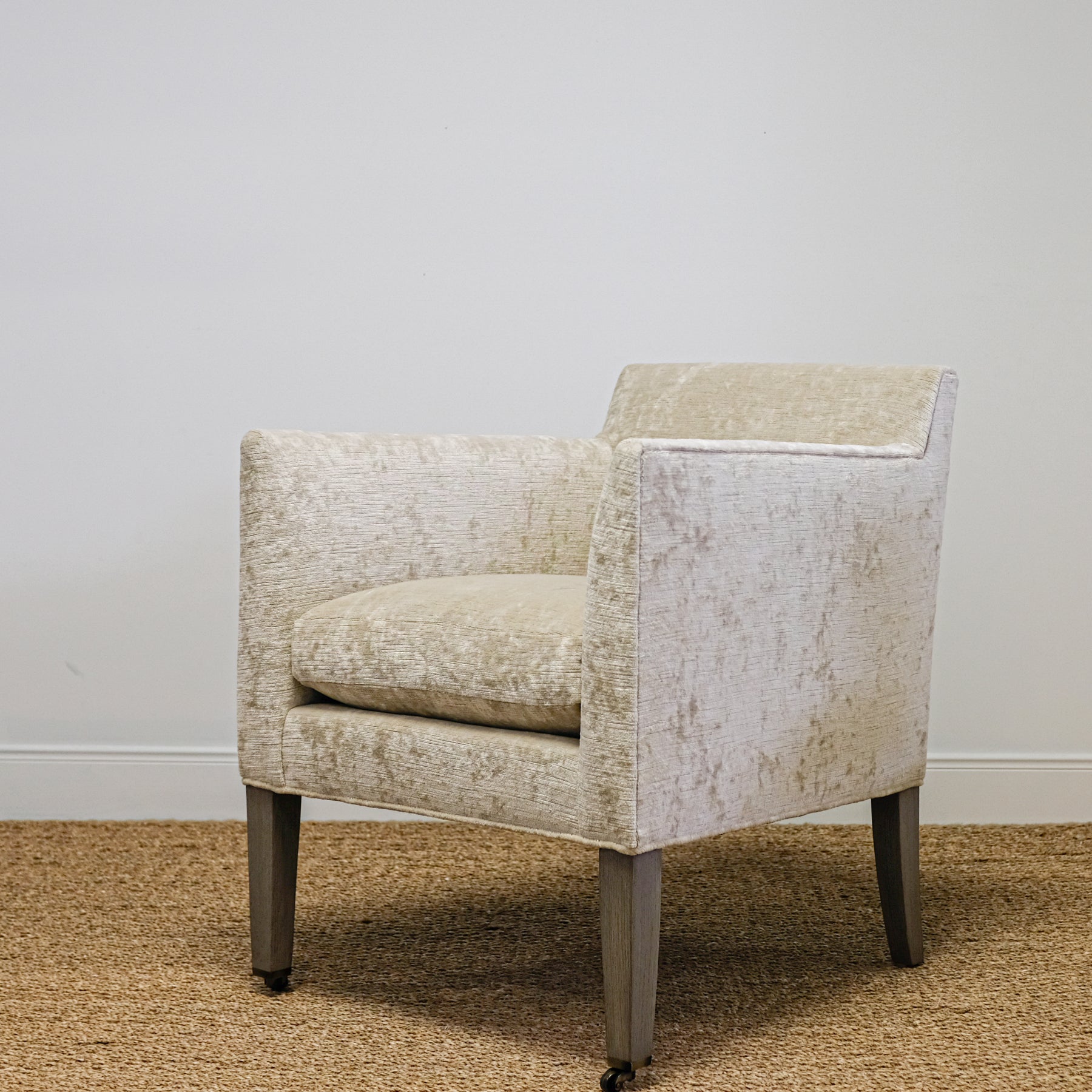 angled view of upholstered chair in mottled beige velvet with exposed wood leg, casters on front legs