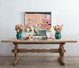 antique dining table set with vases, plates, and decorative objects. Pink abstract art on the wall behind.