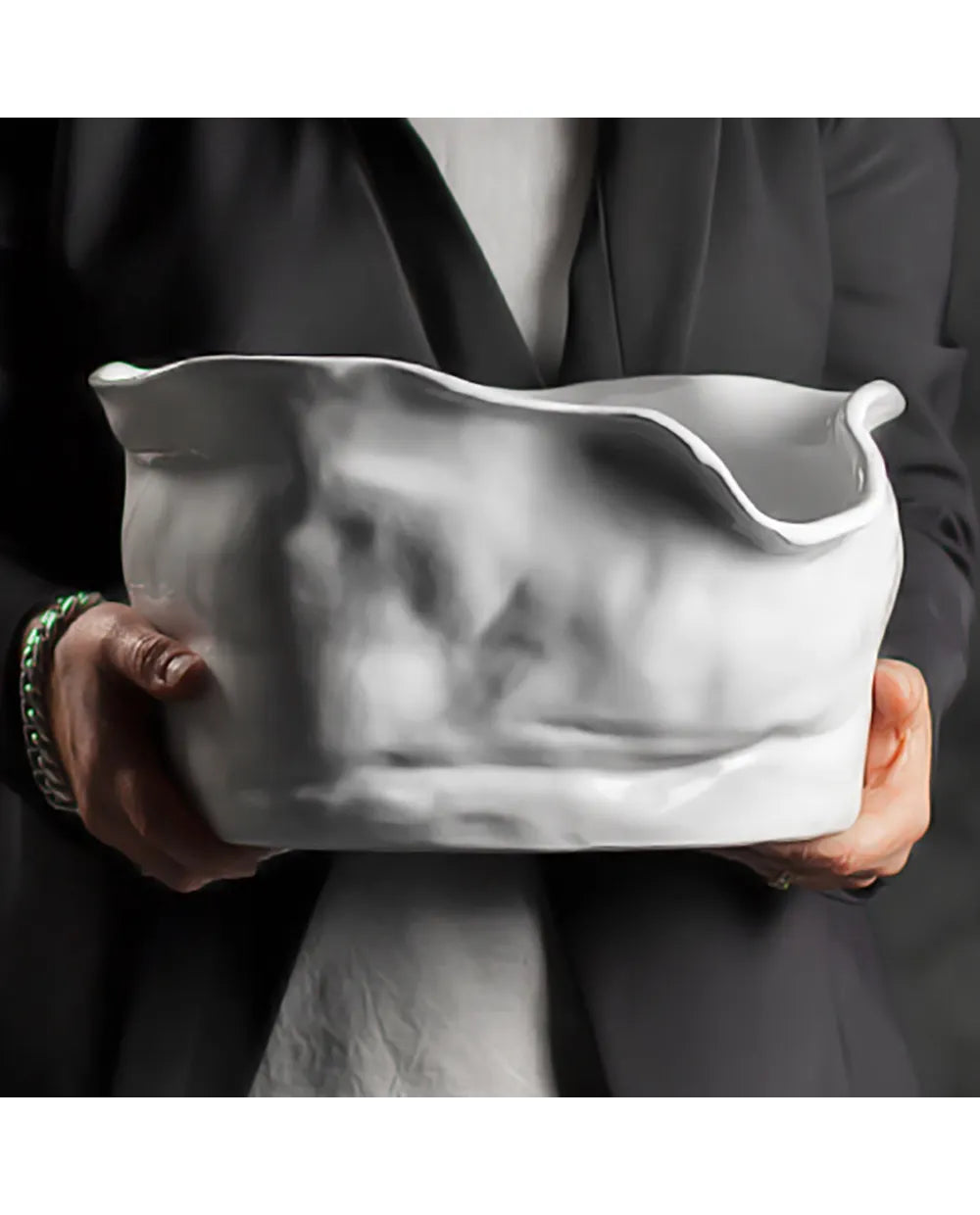 A person in a dark blazer holds a large, sculptural white ceramic vessel with a highly organic, wavy, crumpled surface texture.