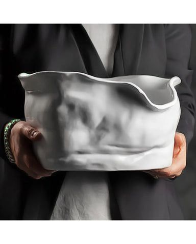 A person in a dark blazer holds a large, sculptural white ceramic vessel with a highly organic, wavy, crumpled surface texture.