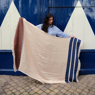 Woman holding a large beige blanket with brown and blue stripes against a blue and white wall.