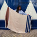 Woman holding a large beige blanket with brown and blue stripes against a blue and white wall.