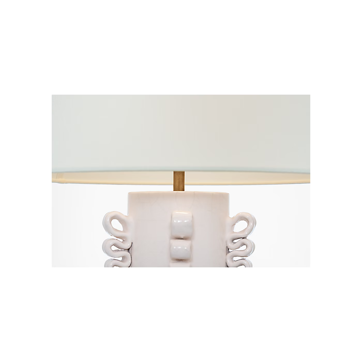 Close-up of a table lamp base with a white ceramic body, sculptural side details, a gold-toned central finial, and a white drum shade.
