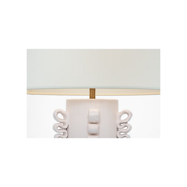 Close-up of a table lamp base with a white ceramic body, sculptural side details, a gold-toned central finial, and a white drum shade.