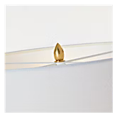 A close-up of a small gold teardrop-shaped crystal or gemstone resting on a clear curved glass surface against a white background.