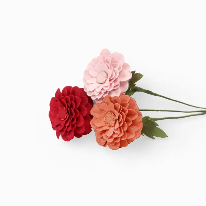 Three artificial dahlia-style flowers on stems against a plain white background: one red bloom, one coral-orange bloom, and one pale pink bloom with green leaves and stems.
