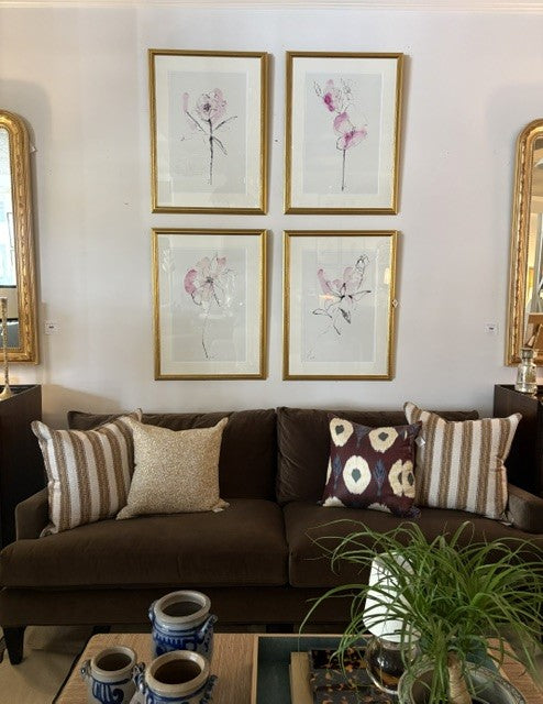 A styled living room scene featuring a dark brown sofa with striped and patterned throw pillows, set against a white wall decorated with four framed botanical prints in gold frames. A wooden coffee table with blue and white ceramic vases and a green potted plant is visible in the foreground.