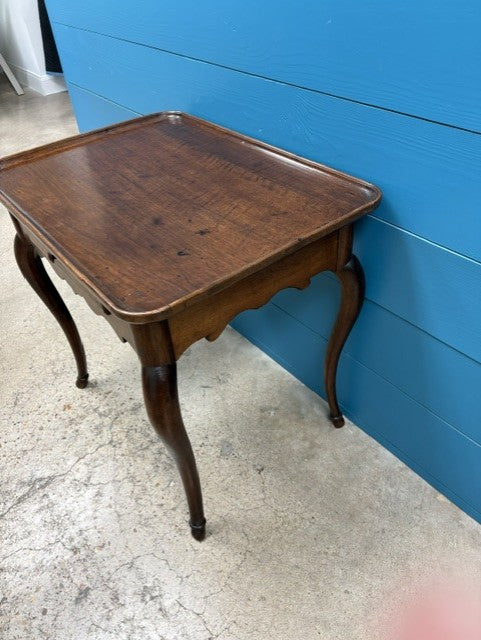 A dark brown wooden side table with curved cabriole-style legs, photographed in a warehouse or showroom setting against a bright blue paneled wall and concrete floor.