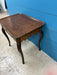 A dark brown wooden side table with curved cabriole-style legs, photographed in a warehouse or showroom setting against a bright blue paneled wall and concrete floor.
