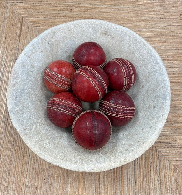 Red cricket balls with white stitching in a stone bowl on a wooden surface