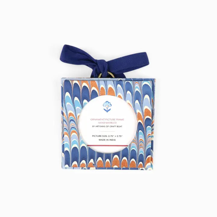 A square ornament picture frame by Artisans of Craft Rug, wrapped in a marbled blue, orange, and white patterned paper and tied with a navy blue ribbon, shown on a plain white background.