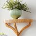 A light natural rattan wall shelf with curved legs, holding a ribbed light green ceramic planter filled with green foliage, set against a plain off-white wall.