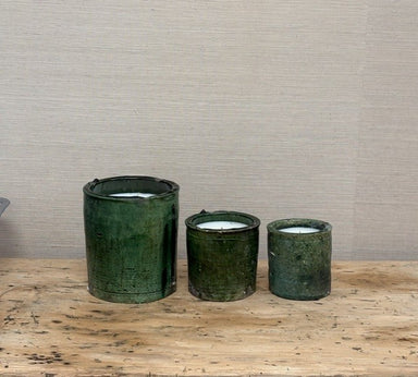 Three dark green ceramic candle holders of varying sizes arranged on a rustic wooden surface against a neutral textured beige wall.