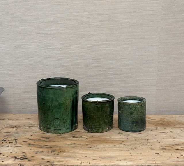Three dark green ceramic candle holders of varying sizes arranged on a rustic wooden surface against a neutral textured beige wall.