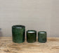 Three dark green ceramic candle holders of varying sizes arranged on a rustic wooden surface against a neutral textured beige wall.
