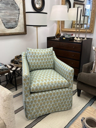 A slipcovered armchair with a light blue and beige geometric pattern, shown in a furnished room with a dark wood dresser, lamps, and other home decor.