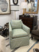 A slipcovered armchair with a light blue and beige geometric pattern, shown in a furnished room with a dark wood dresser, lamps, and other home decor.