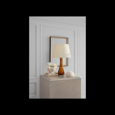 A table lamp with a warm amber glass base and a cream fabric drum shade, styled on a beige pedestal against a white paneled wall with a gold-framed mirror behind it.