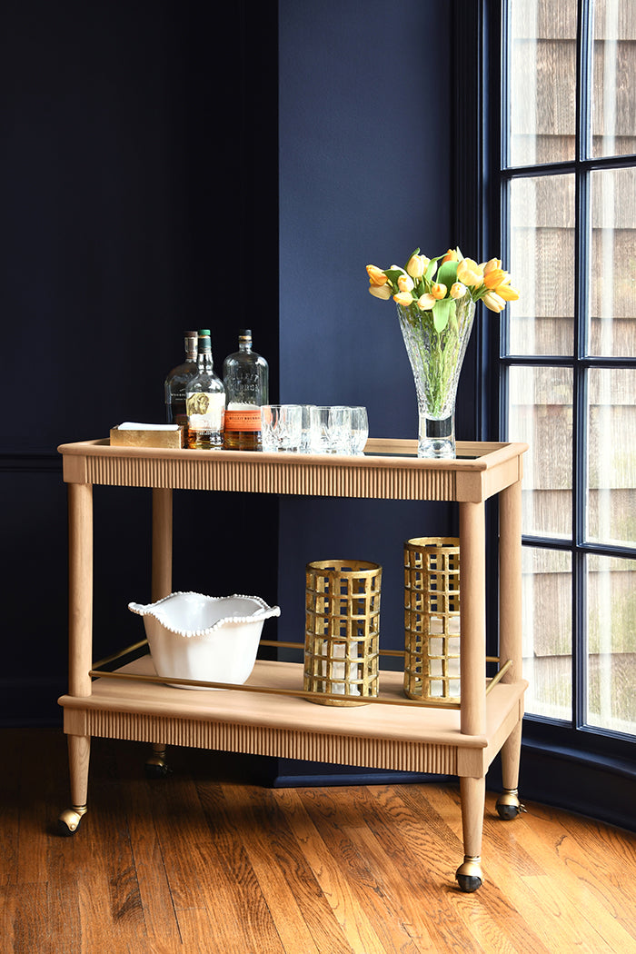 Wooden bar cart with drinks and decor against a dark wall with a window.