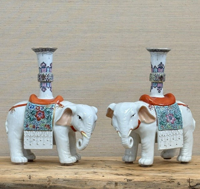 A pair of white porcelain elephant candlestick holders with colorful floral patterns and orange-brown saddle details, displayed on a wooden surface against a neutral textured background.