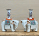 A pair of white porcelain elephant candlestick holders with colorful floral patterns and orange-brown saddle details, displayed on a wooden surface against a neutral textured background.