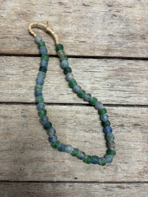 A beaded necklace made of round, faceted beads in shades of green, blue, and gray, strung on a tan cord and laid out on a rustic wooden surface.