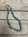 A beaded necklace made of round, faceted beads in shades of green, blue, and gray, strung on a tan cord and laid out on a rustic wooden surface.