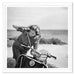A framed black and white photograph print showing a woman leaning on a scooter outdoors near a beach or open landscape. The print is presented in a white frame with a white mat border.