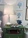 Green nightstand with lamp, decorative items, and framed floral art on a wall.