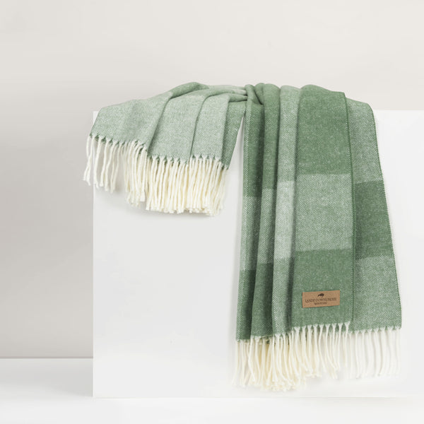 A green and white checkered blanket with cream-colored fringe, draped over a white display block. A small brown leather brand label is visible near the bottom right corner.
