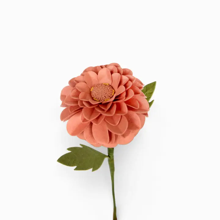 A single artificial flower with layered peach-pink petals, a yellow-orange center, and green leaves on a stem, photographed against a plain white background.