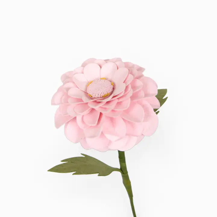 A single artificial pink flower with layered petals, a yellow-tinted center, and green leaves on a stem, photographed against a plain white background.