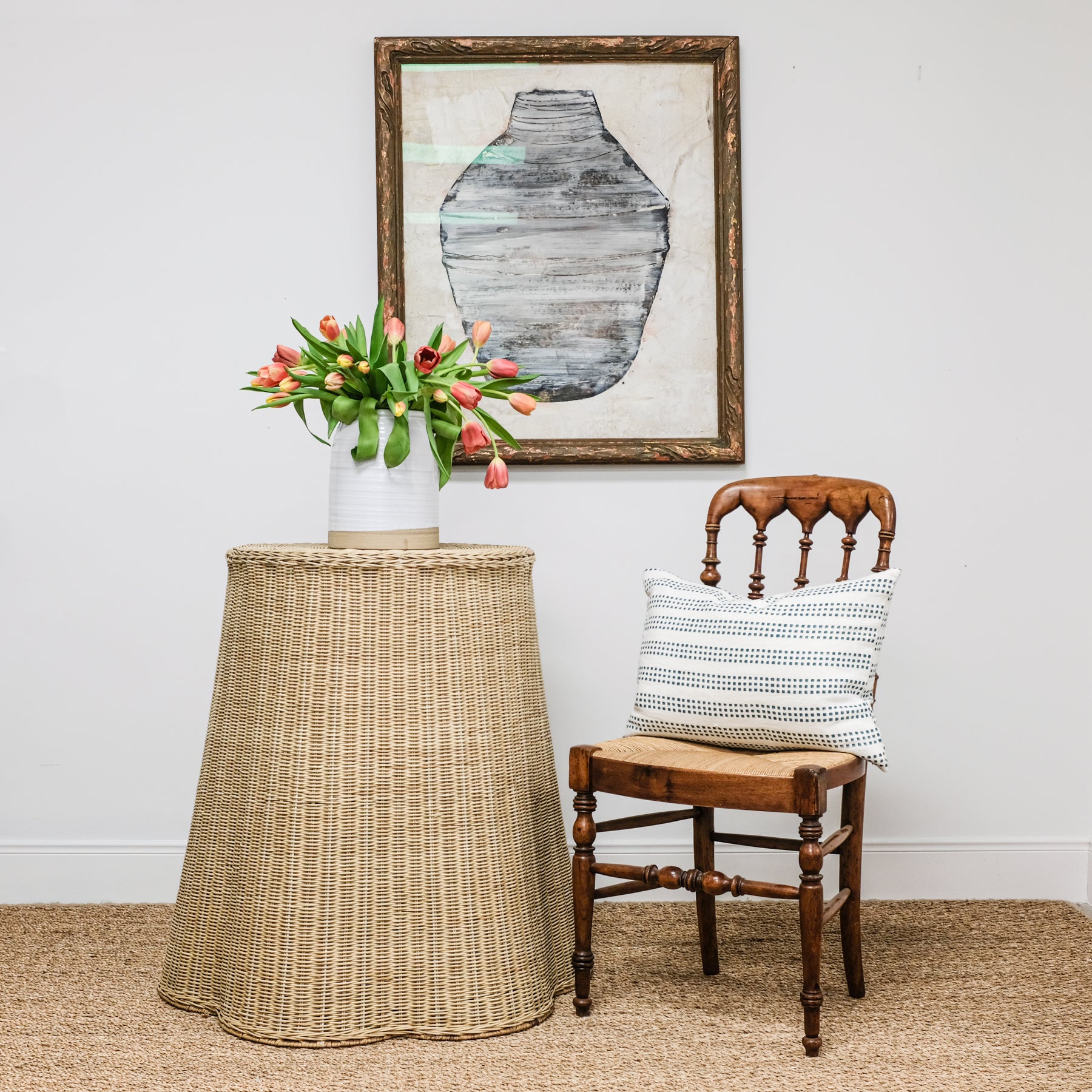 scalloped wicker side table with chair pillow and art. vase of tulips