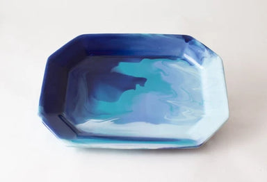 An octagonal shallow dish with a marbled resin design featuring shades of blue, teal, and white, photographed on a plain off-white background.