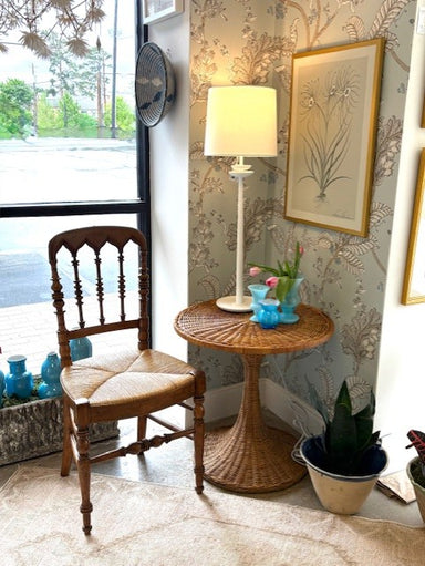 Small table with lamp and decorative items next to a wooden chair in a room with floral wallpaper.